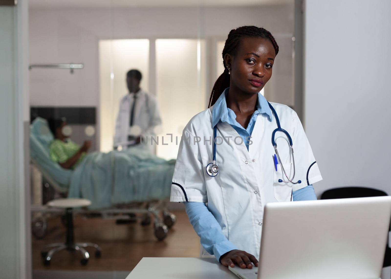 African american doctor looking at camera in hospital ward with medical equipment and instruments. Portrait of african ethnicity woman with stethoscope and white coat at clinic desk