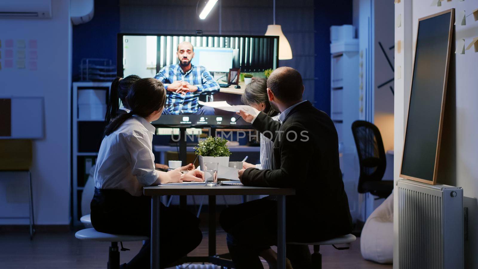 Multi ethnic businesspeople discussing with remote entrepreneur in wheelchair during online videocall conference at night in company meeting office room. Diverse focused teamwork brainstorming ideas