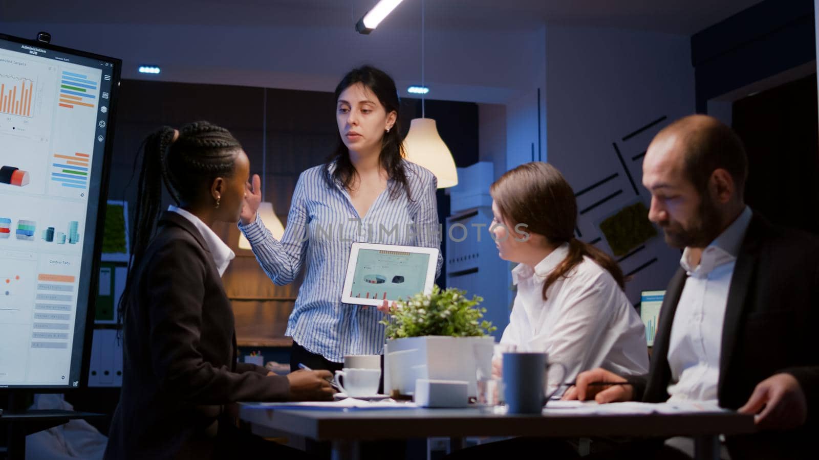Overworked workaholic entrepreneur woman shwoing marketing graphs using tablet overworking at company solution late at night in meeting room. Diverse multi-ethnic teamwork solving financial strategy