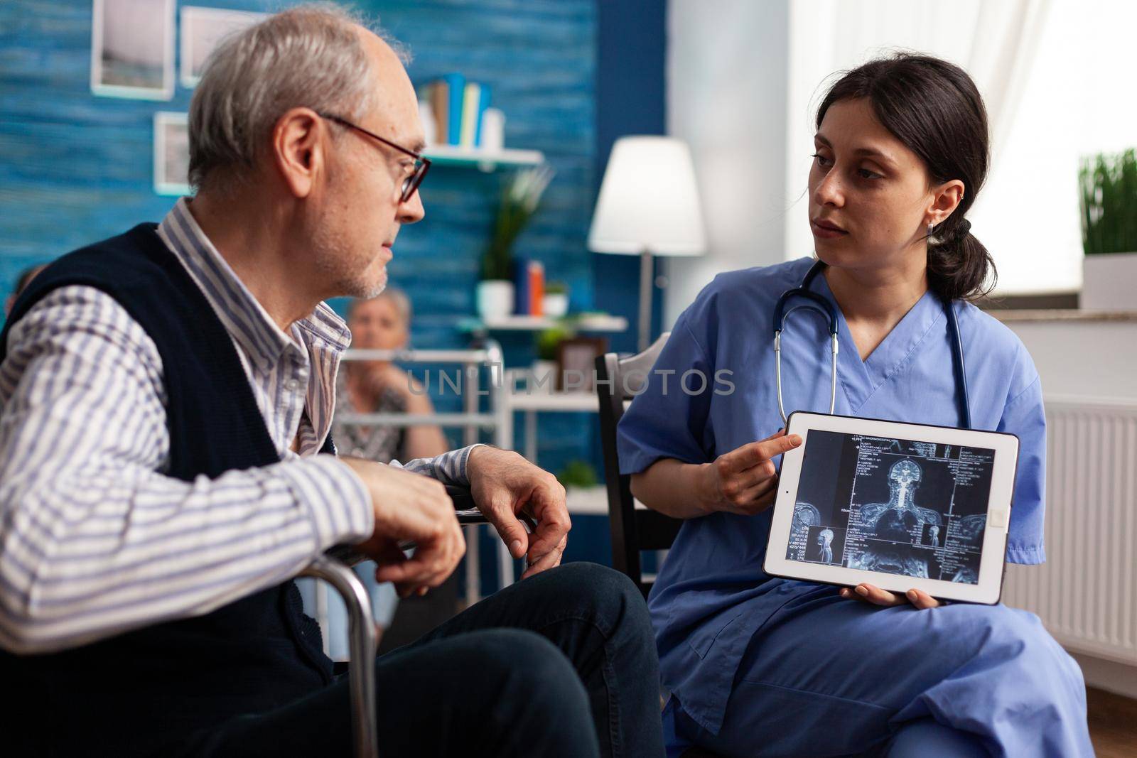 Social nurse woman holding tablet with radiography on display explaining medical treatment to disabled senior man. Social services nursing elderly retired male. Healthcare assistance