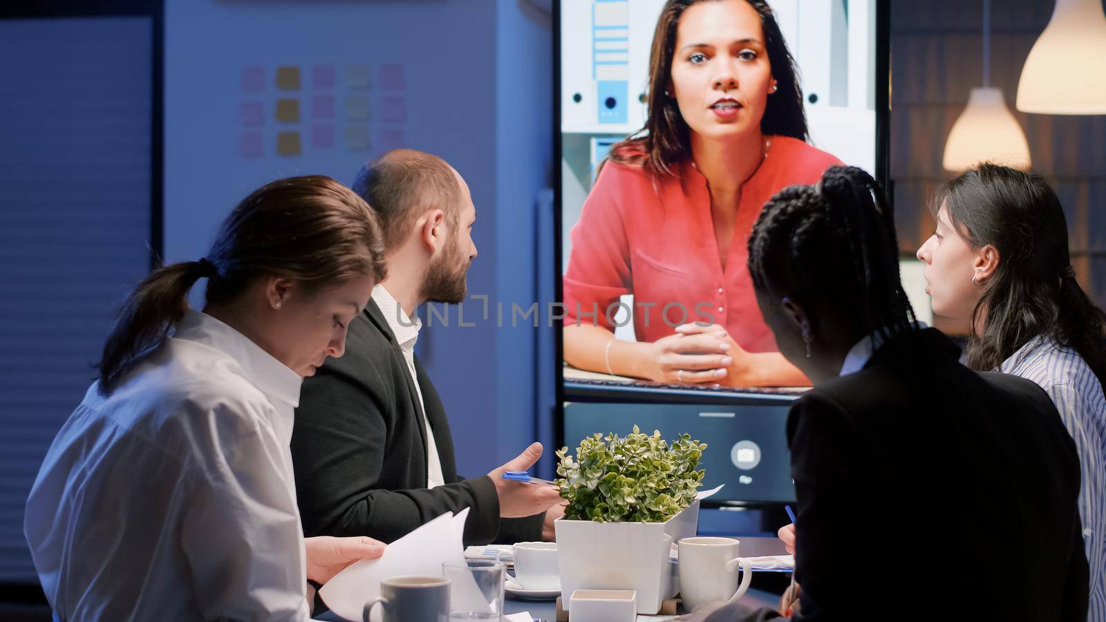 Diverse multi-ethnic businesspeople discussing with remote manager during online videocall conference meeting late at night in company office room. Focused teamwork brainstorming ideas