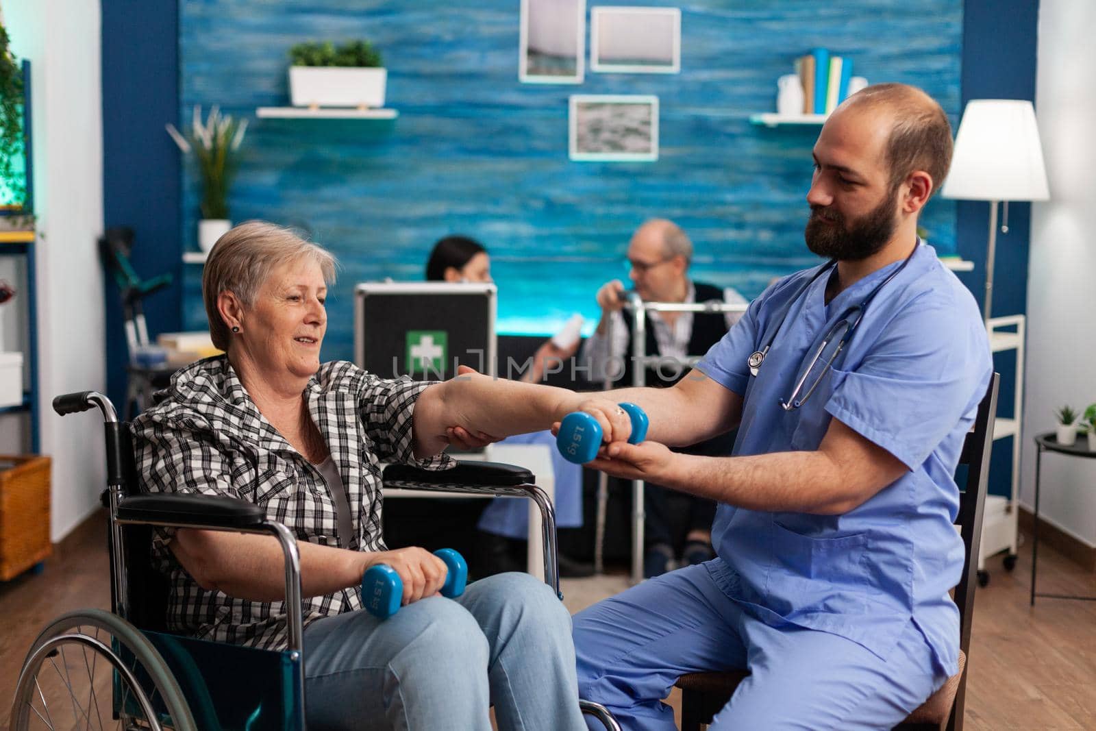 Male nurse helping senior retired disabled woman in wheelchair to rehabilitate using dumbbels during recovery session in nursing home. Elderly woman exercising physiotherapy with social worker