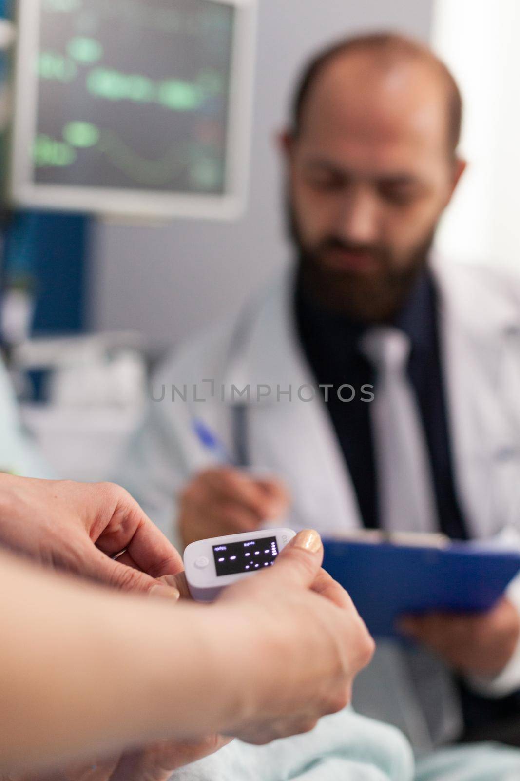 Asisstant nurse puts medical oximeter on finger while doctor man writes heartbeat puls rate on clipbord during consultation. Social services nursing elderly retired female. Healthcare assistance