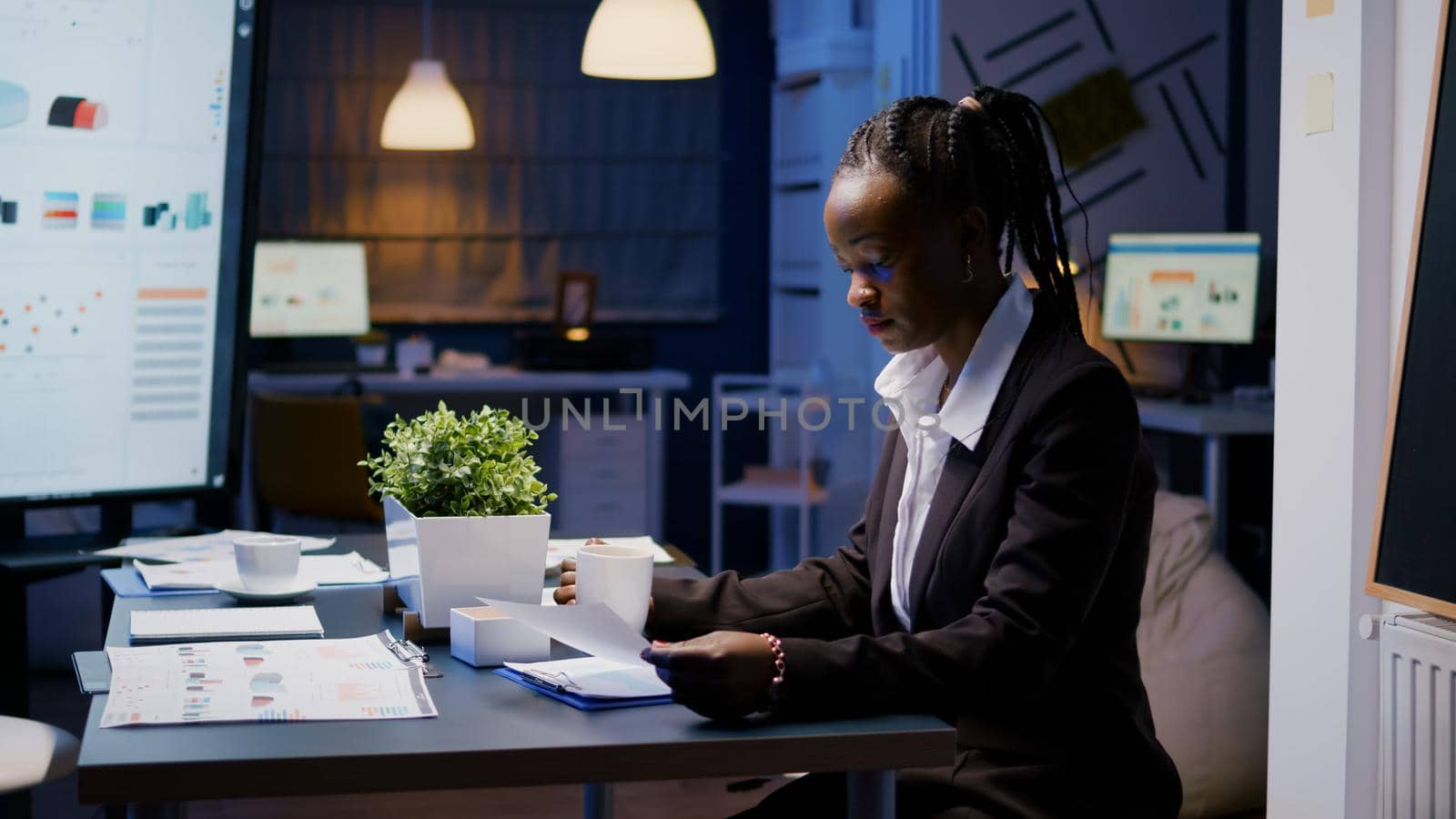 Workaholic african american businesswoman analysing marketing statistics on paperwork drinking coffee sitting at table late at night. Business manager with dark skin working in office meeting room