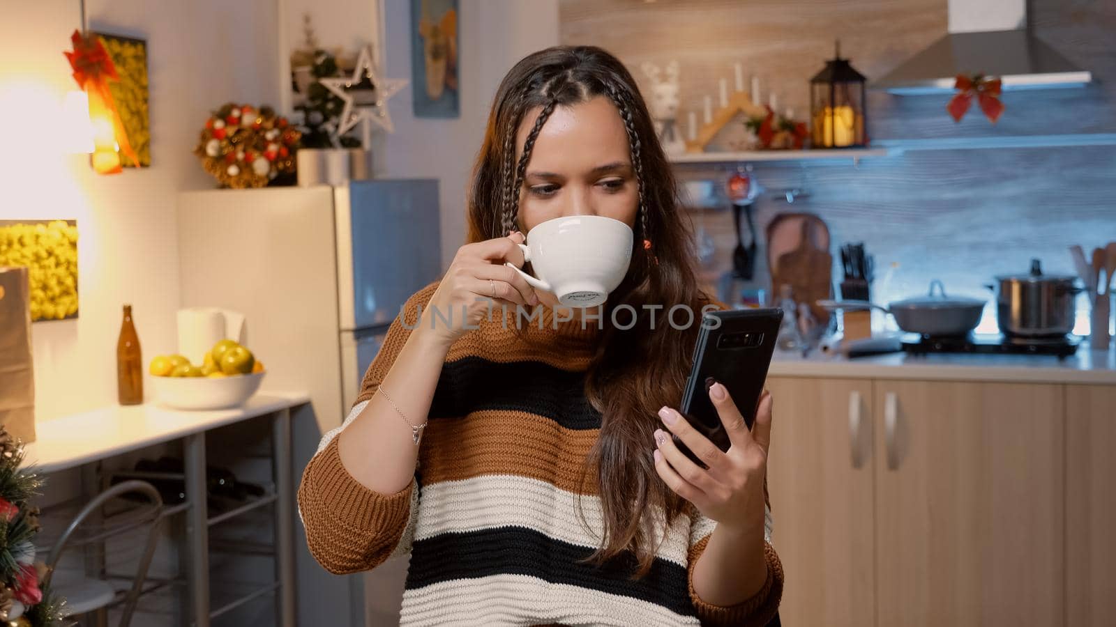 Caucasian adult using video call modern technology on smartphone in kitchen with christmas decorations and ornaments. Woman talking with friends on internet about holiday celebration