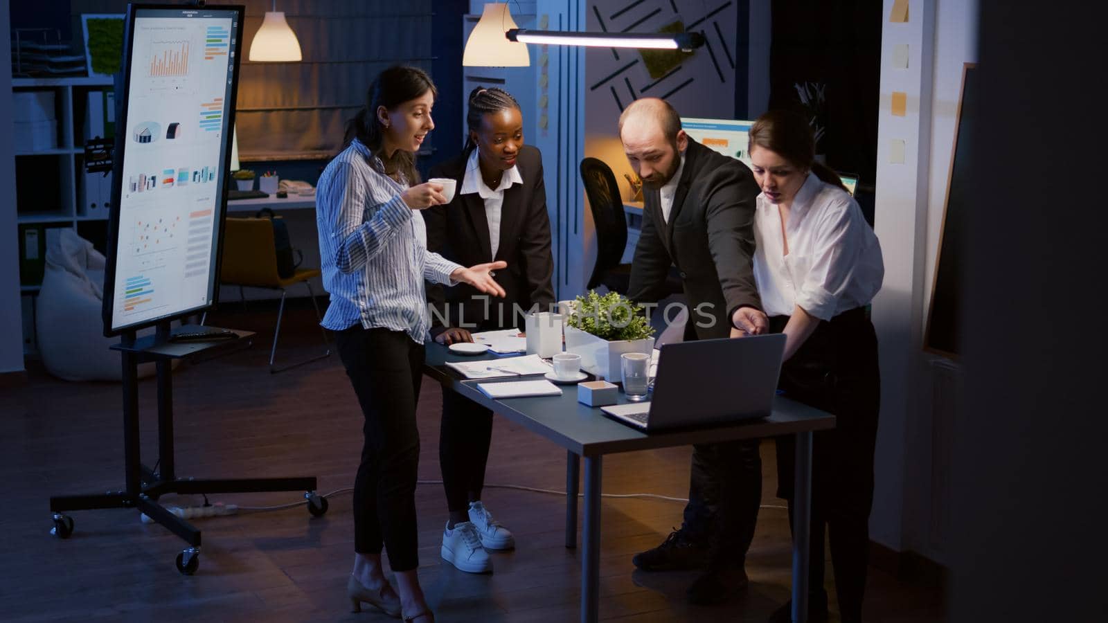 Excited multi ethnic businesspeople receiving good news clapping while standing at table conference overworking in office meeting room late at night. Diverse teamwork looking at company presentation