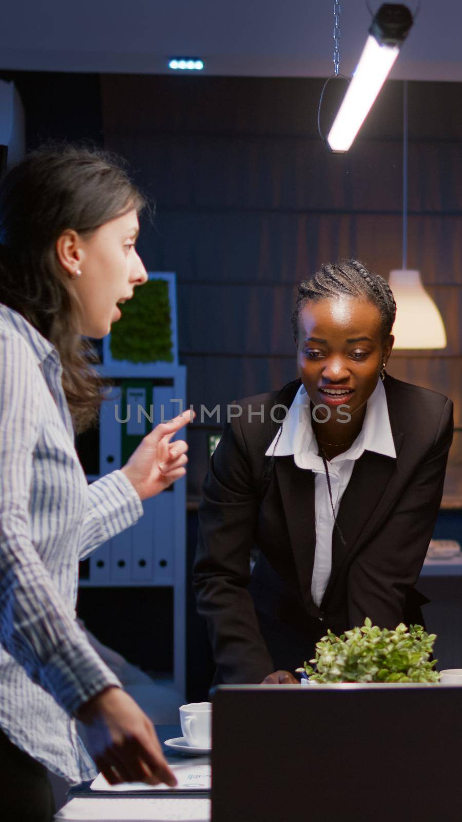 Diverse multi ethnic teamwork brainstorming company ideas analyzing financial graphs presentation using laptop computer. Businesspeople overworking in meeting room late at night discussing solution