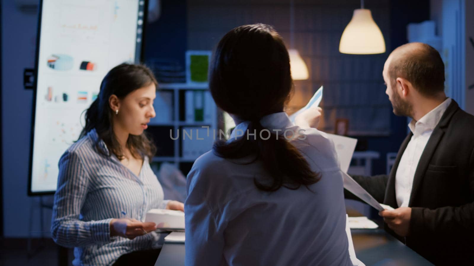 Multi ethnic managers teamwork checking financial graphs analyzing business paperwork working in meeting room late at night. Diverse businesspeople solving company ideas for management presentation