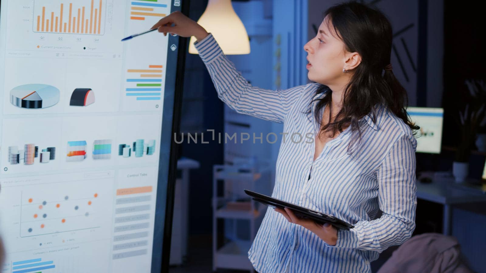 Workaholic focused businesswoman explaining management solution pointing strategy on monitor overworking in company business office meeting room. Multi-ethnic coworkers discussing ideas in evening