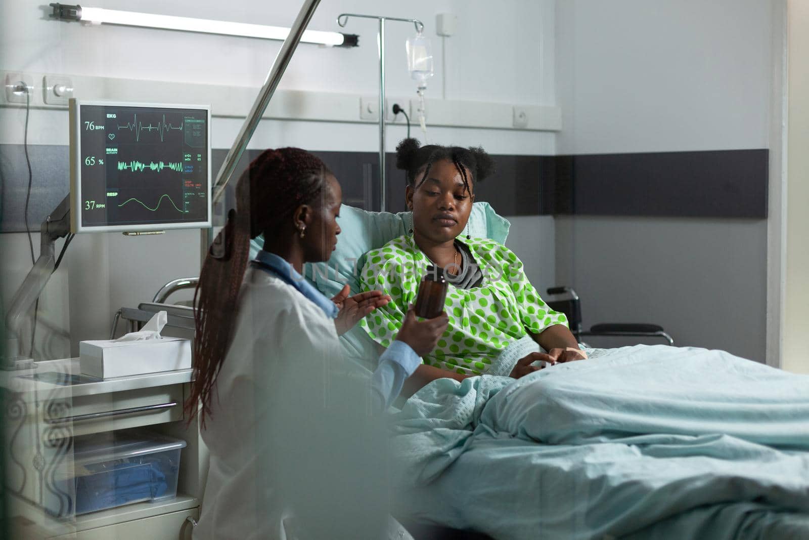 African american people talking about treatment in hospital ward. Professional doctor showing bottle of pills, drugs, prescription medicine to young patient with illness sitting in bed