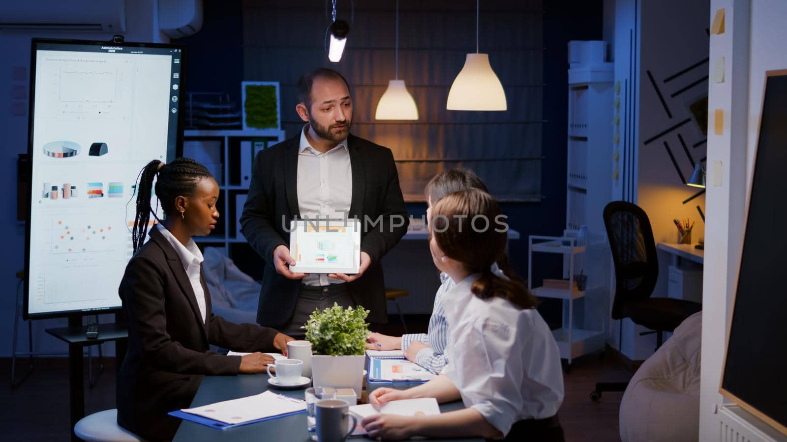 Focused workaholic entrepreneur man working overtime presenting company statistics using tablet. Diverse multi-ethnic businesspeople overworking in office meeting room late at night