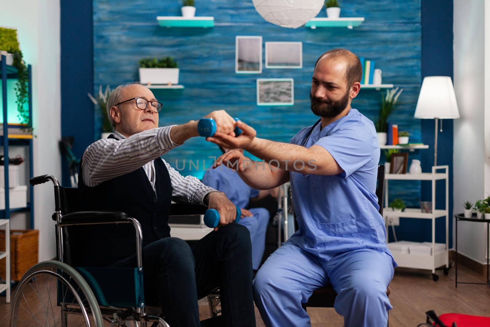 Practitioner man doctor helping retiree senior male in wheelchair to do physiotherapy strength exercise using dumbbell. Social nursing services at home. Healthcare recovery. Support worker helper