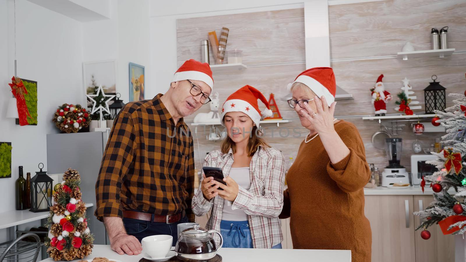 Happy family with santa hat greeting remote friends during online videocall meeting using phone celebrating christmas holiday together. Granddaughter enjoying winter season in xmas decorated kitchen