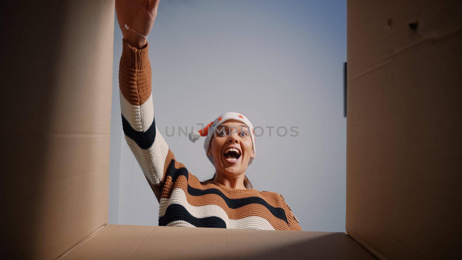 Caucasian woman with santa hat opening present box for surprise at home. Young festive adult smiling looking inside package. Person receiving christmas gift from friends and relatives