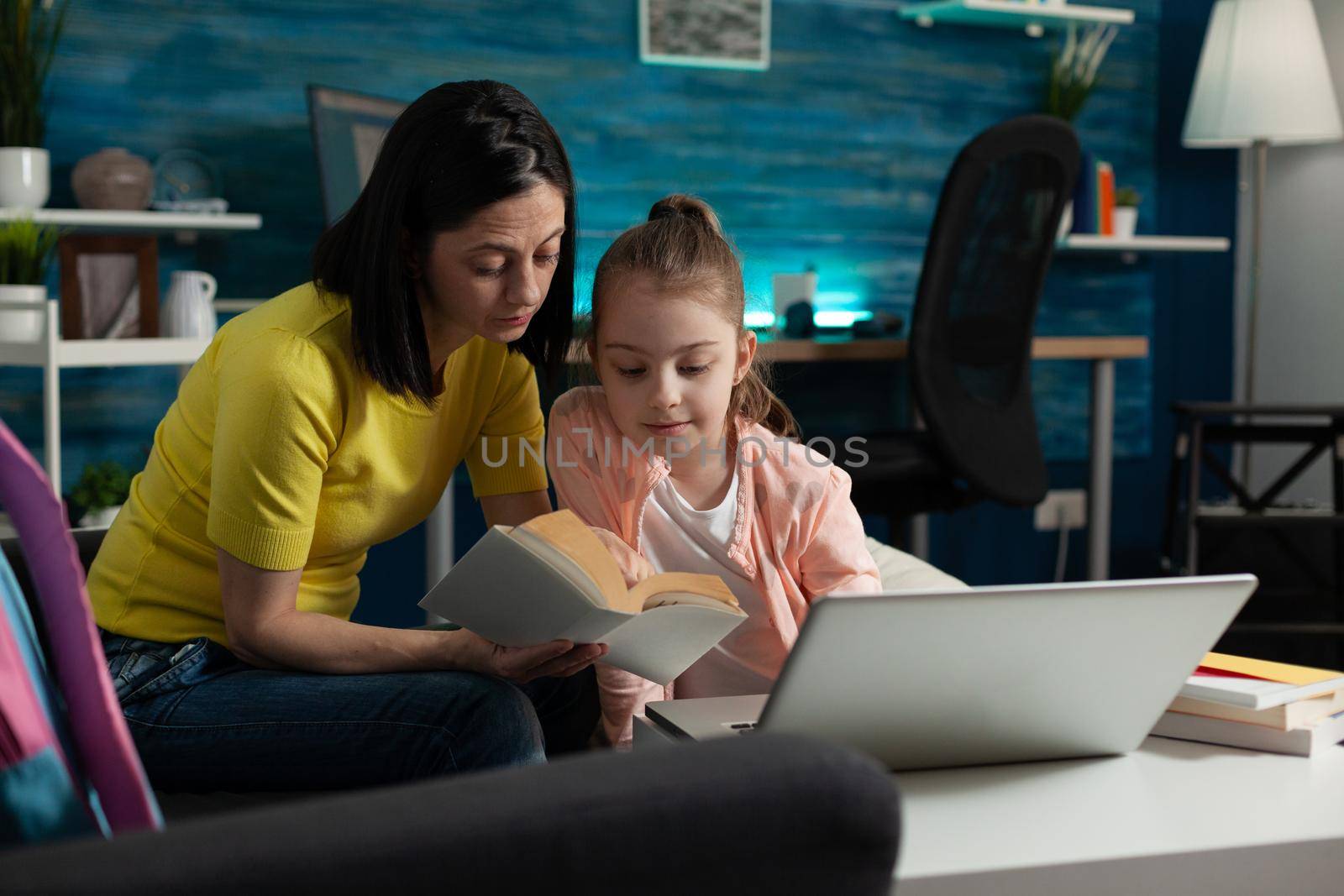 Mother assisting little child on reading school book while sitting at home. Smart girl with modern laptop on desk learning to read and studying for online classroom lesson lecture education