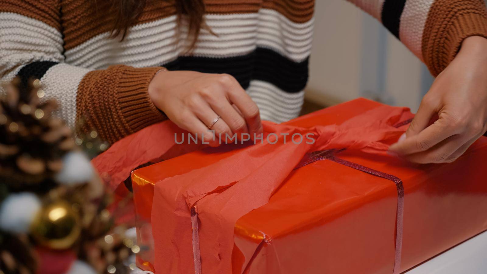 Young adult wrapping gift box in red paper to give to friends at christmas eve celebration party. Woman decorating present with ribbon for winter season joyful tradition in december