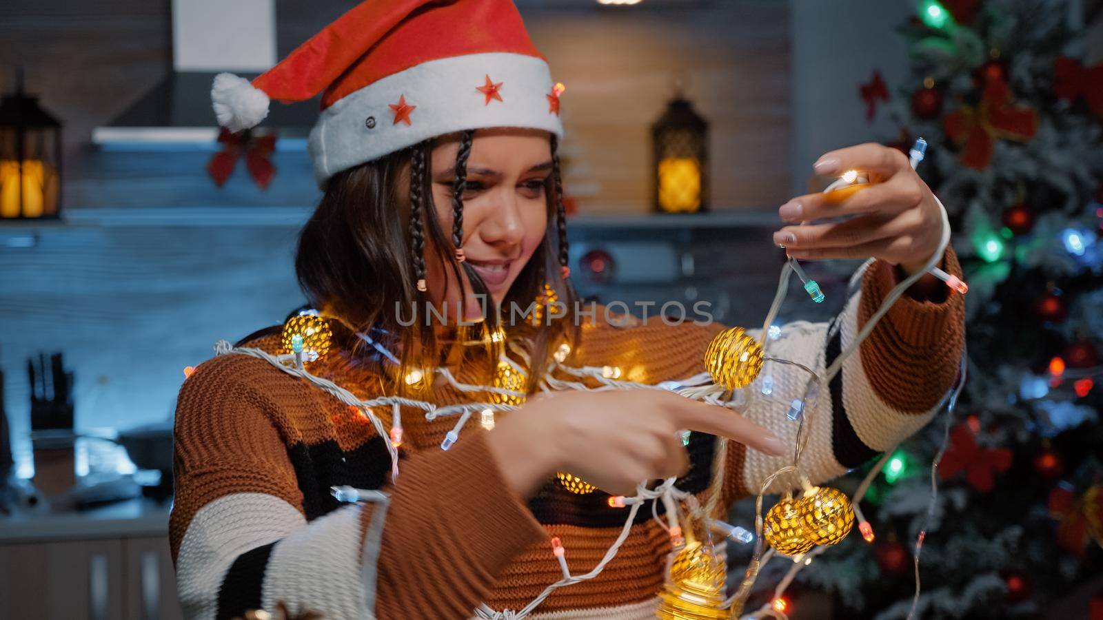 Cheerful woman laughing at tangled rope with lights for christmas decorating. Young adult using ornaments and garland preparing for holiday festivity and december celebration party