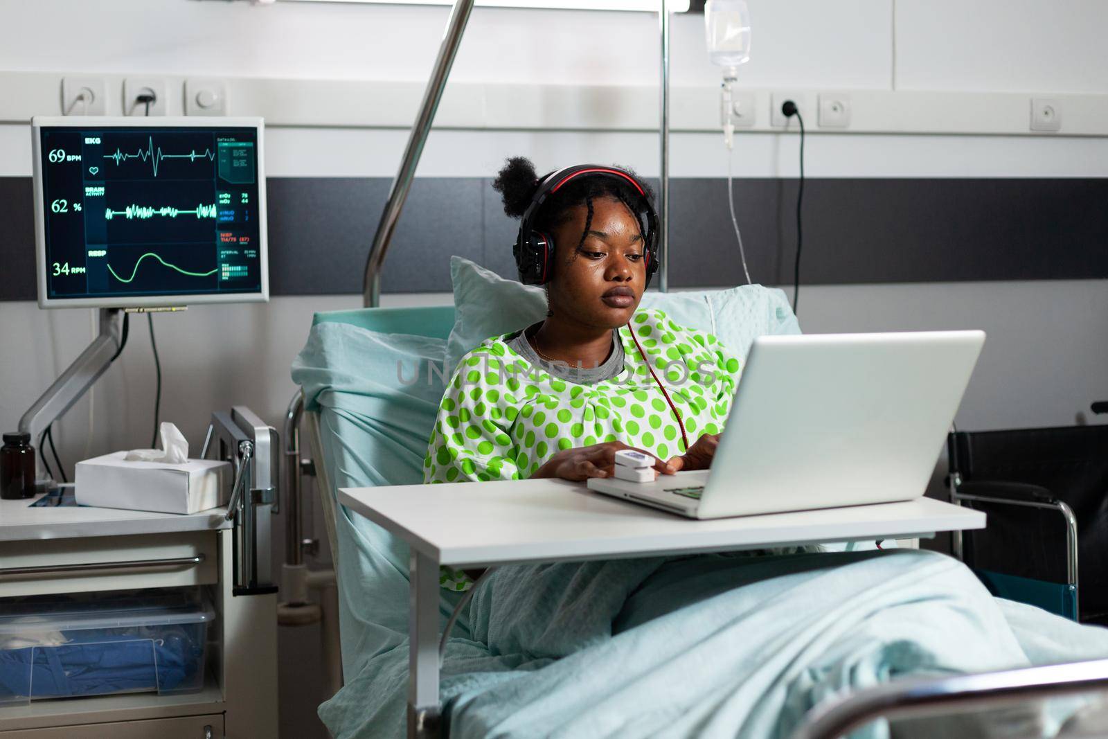 Sick young patient with headphones using laptop in hospital ward. African american teenager with illness, disease recovering in bed while listening to music on online internet technology