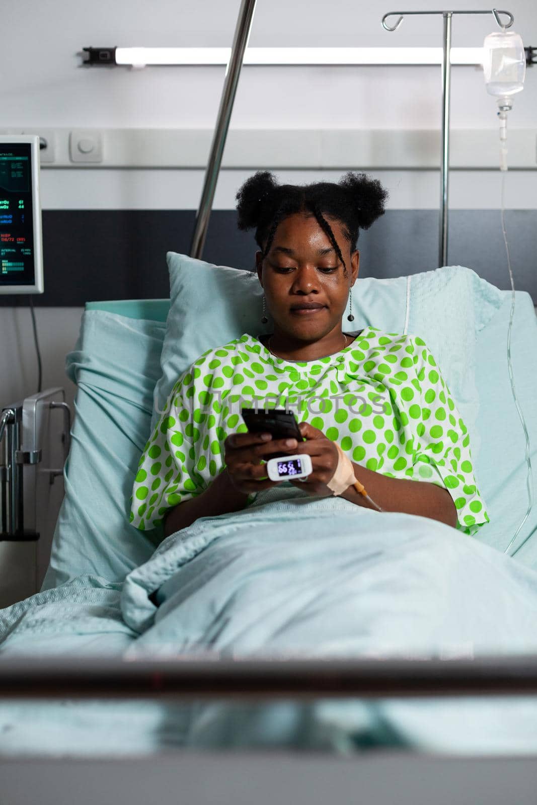 Young african american adult sitting in hospital ward bed using smartphone for web surfing and communication. Teenager patient waiting on medicine and consultation while having online gadget