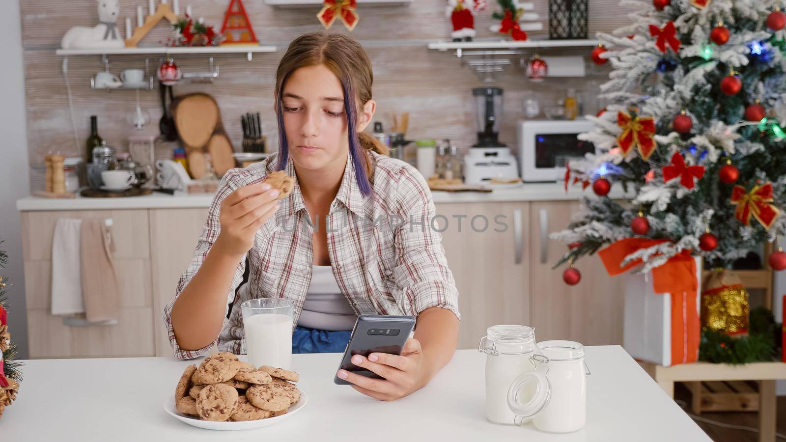 Happy girl enjoying winter holiday sitting at table in xmas decorated kitchen browsing on smartphone watching festive video using phone. Girl celebrating christmas season eating baked cookies