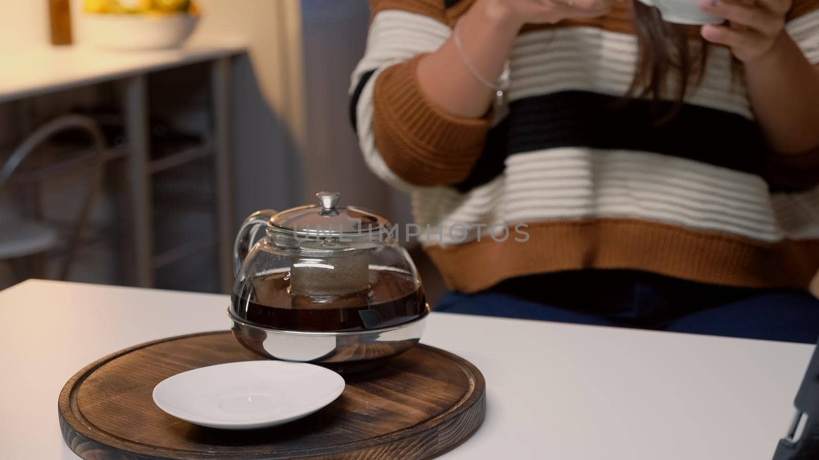 Cup of tea and kettle sitting on white kitchen table at home. Caucasian woman with sweater using tablet for video call while drinking hot winter beverage from mug waiting for holiday dinner