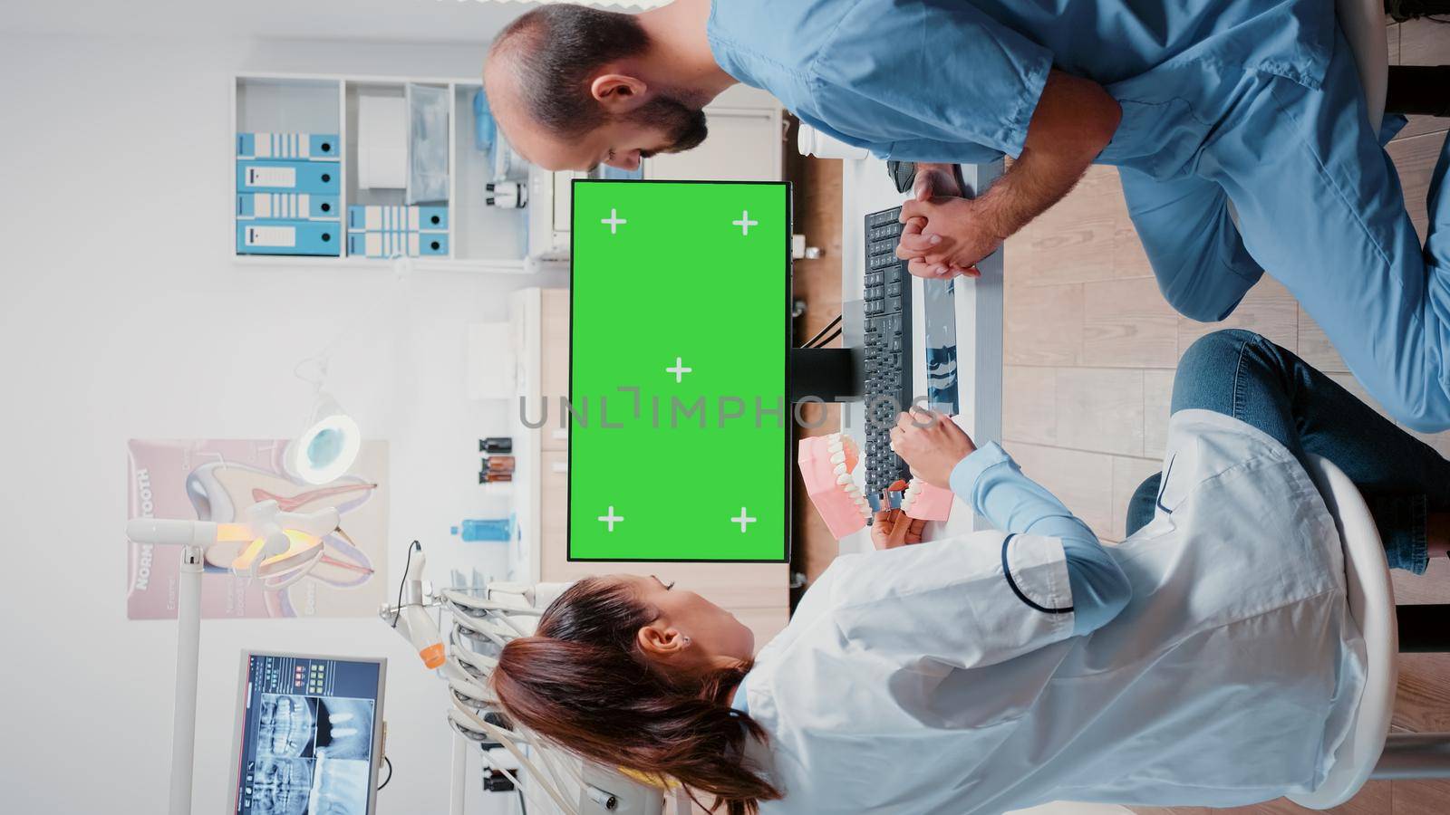 Vertical video: Dentist and assistant looking at green screen on computer for teethcare examination. Man and woman working with dental tools and chroma key with mockup template for oral care.