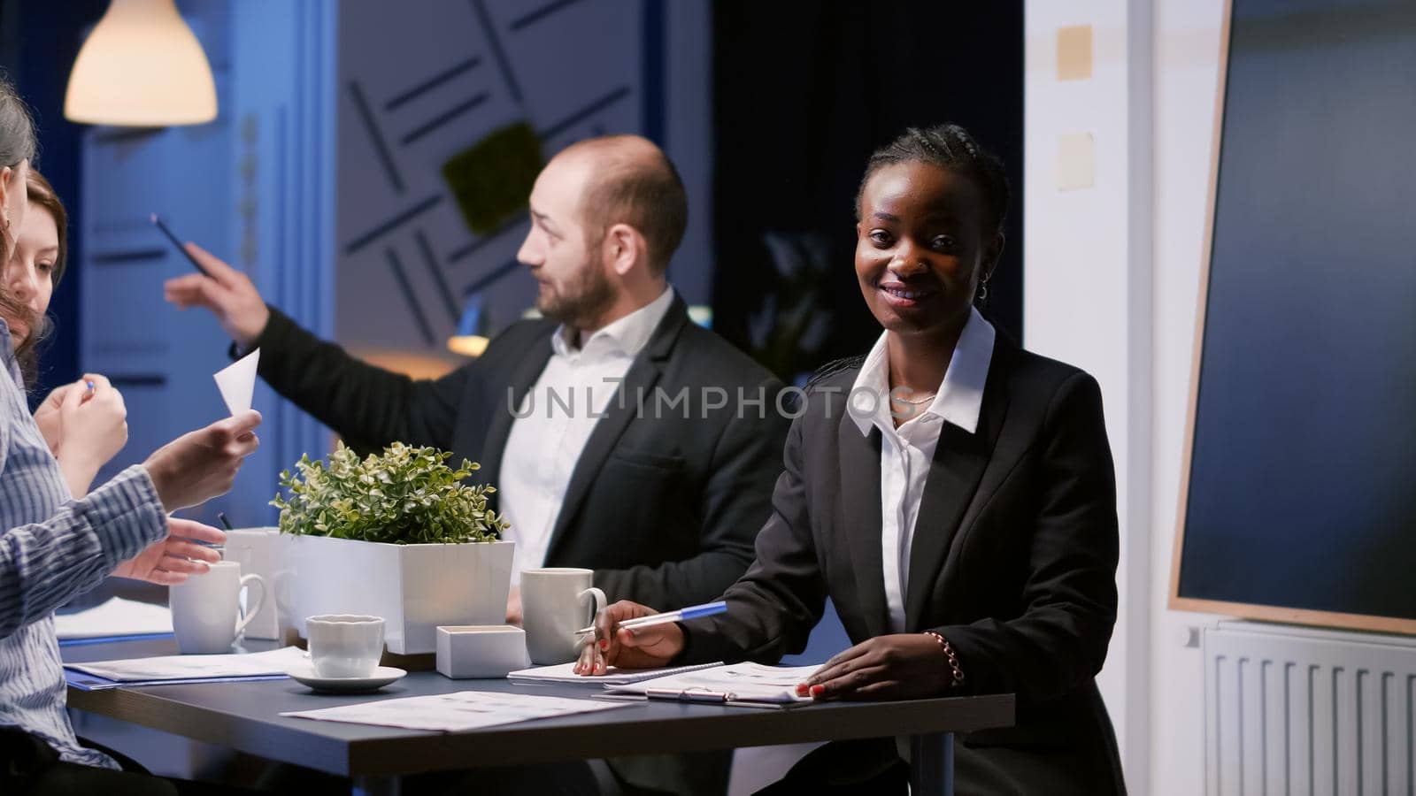 Portrait of focused smiling african american woman sitting at conference table in meeting room working at management investment. Diverse multi-ethnic team discussing company strategy late at night