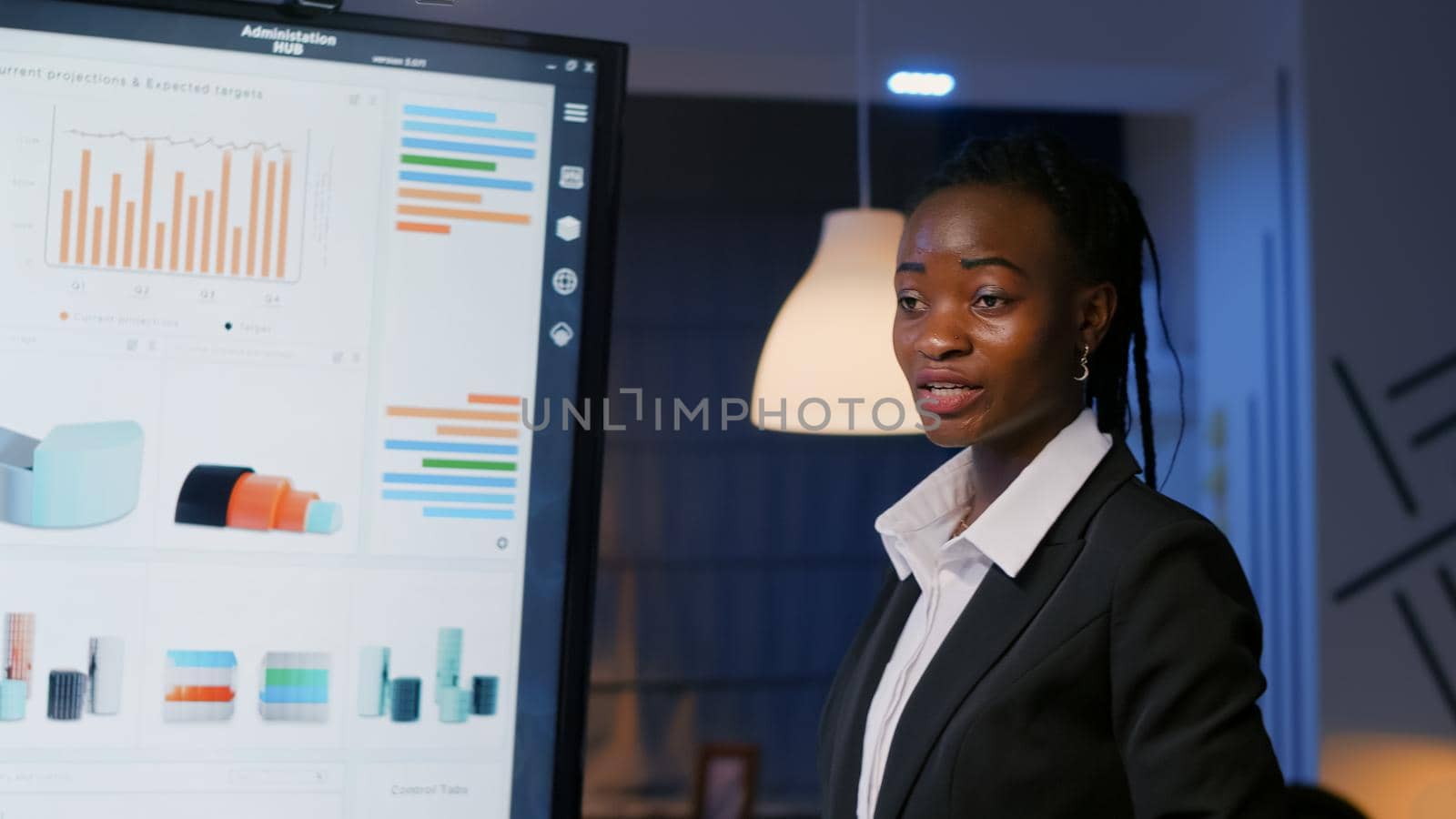 African american executive manager standing in front of presentation monitor analyzing marketing statistics late at night in meeting room. Diverse multi ethnic teamwork brainstorming business ideas