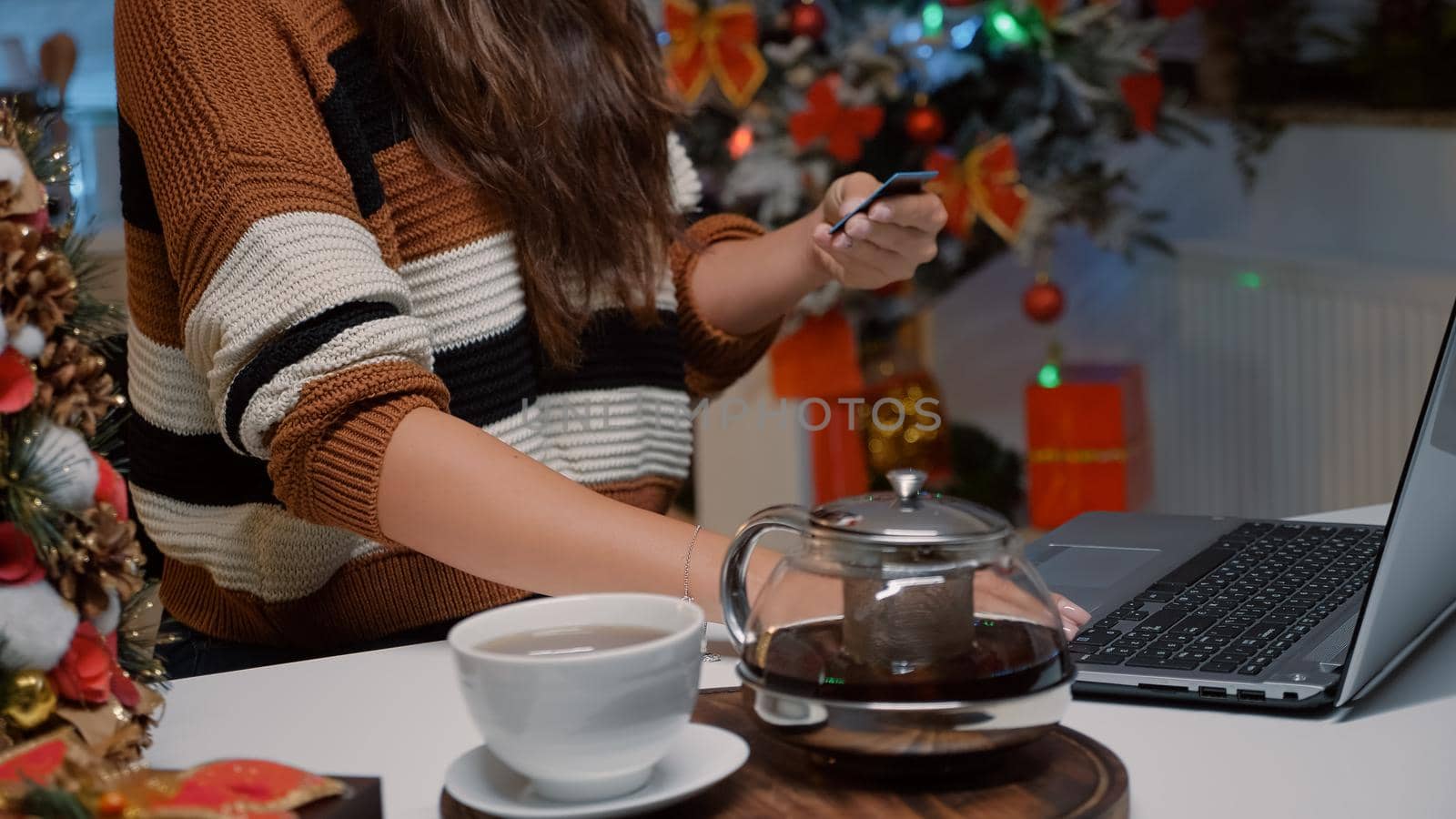 Woman typing credit card number on laptop to shop presents online for friends in christmas decorated kitchen at home. Young person using technology to buy gifts for dinner celebration