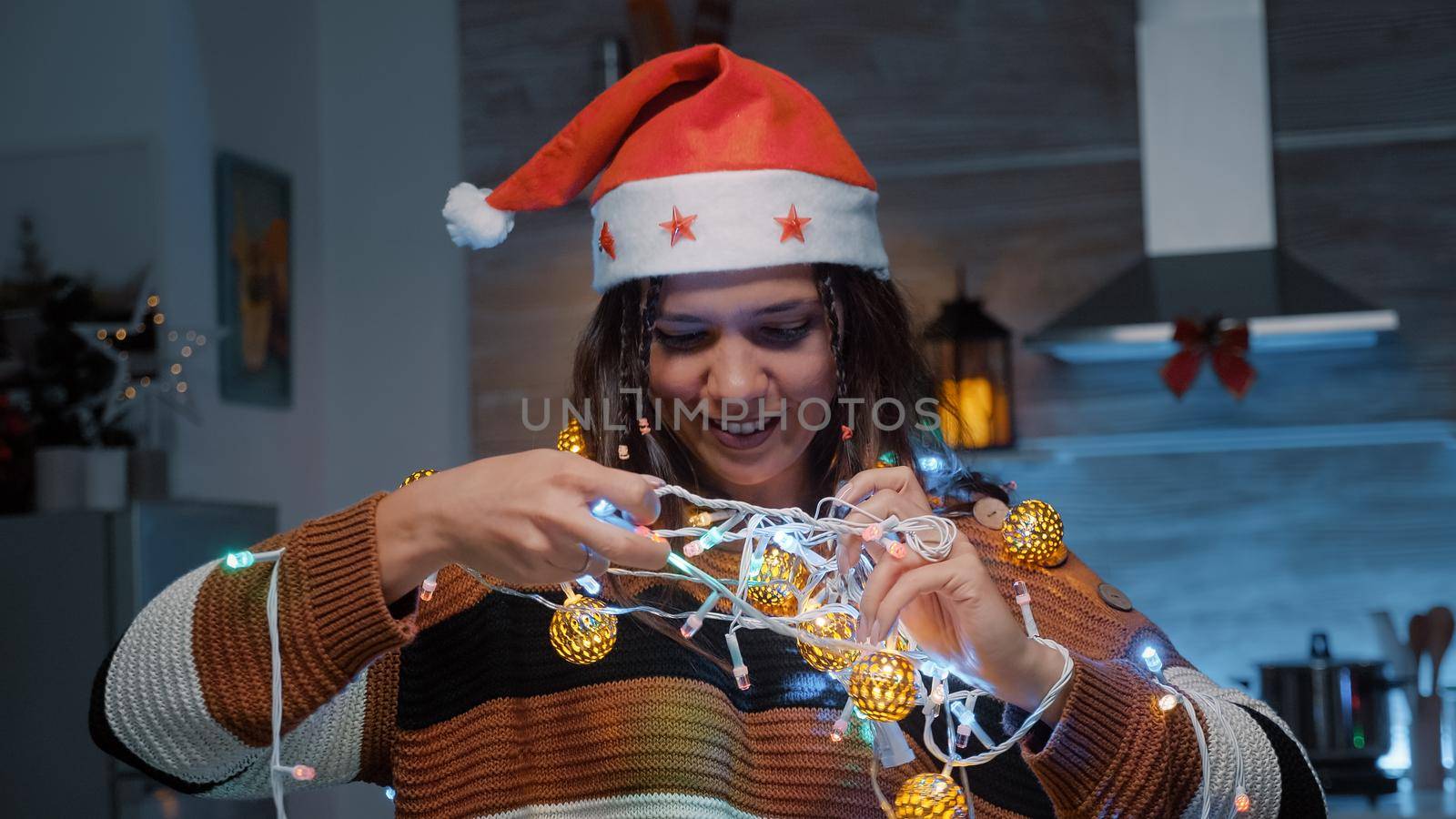 Festive woman with santa hat trying to untangle lights while decorating kitchen for christmas eve festivity. Young adult holding knotted garland for tree and preparing for dinner celebration