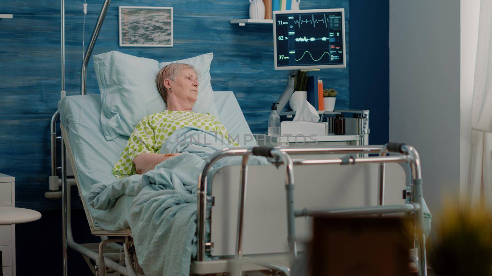 Elder patient with sickness laying in hospital bed with heart rate monitor and IV drip bag for medical assistance and treatment, recovering after surgery. Retired woman resting in nursing home.
