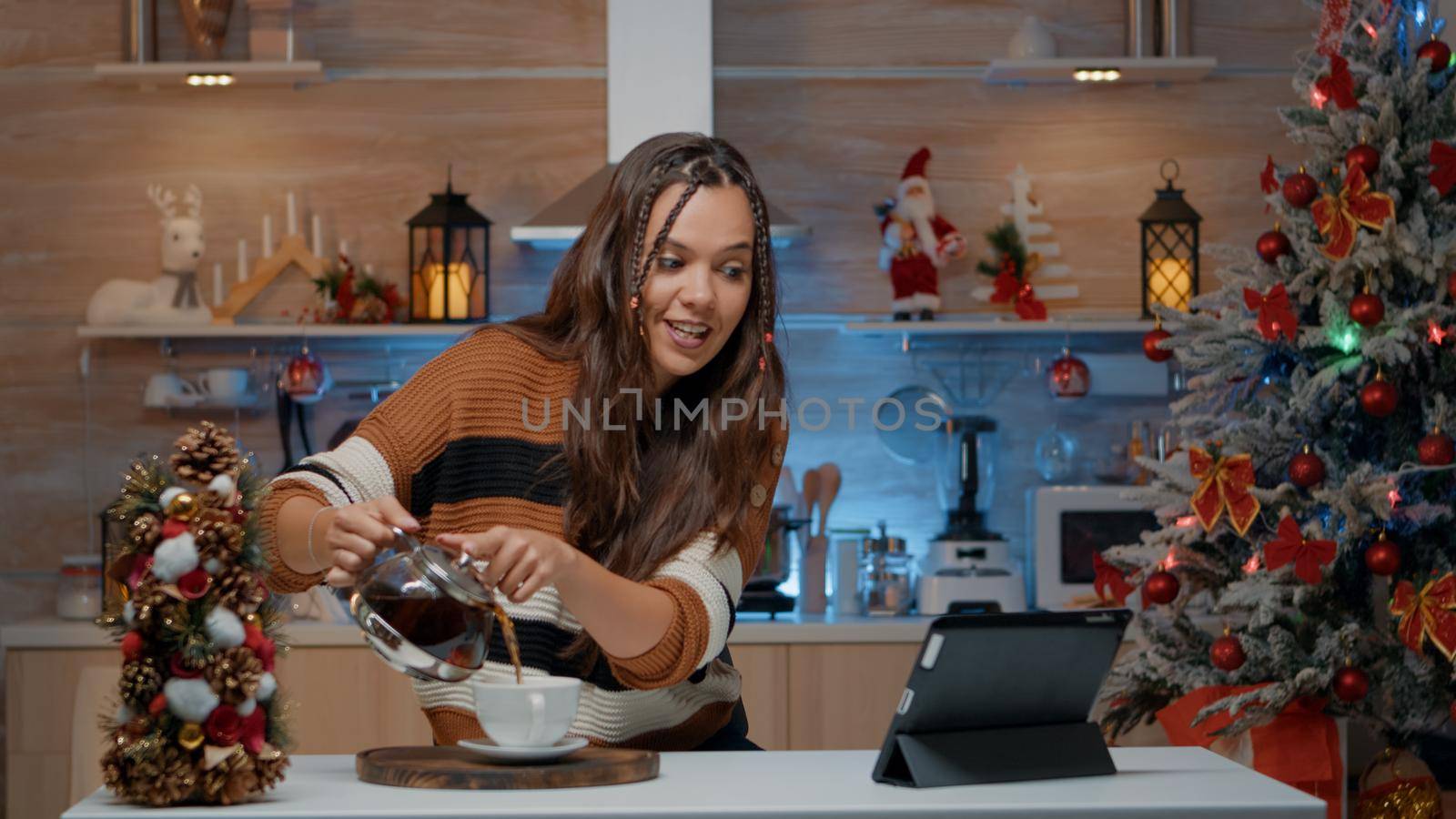 Woman pouring coffee while chatting on video call using tablet. Young adult preparing for festive christmas celebration in kitchen decorated with winter ornaments for seasonal festivity