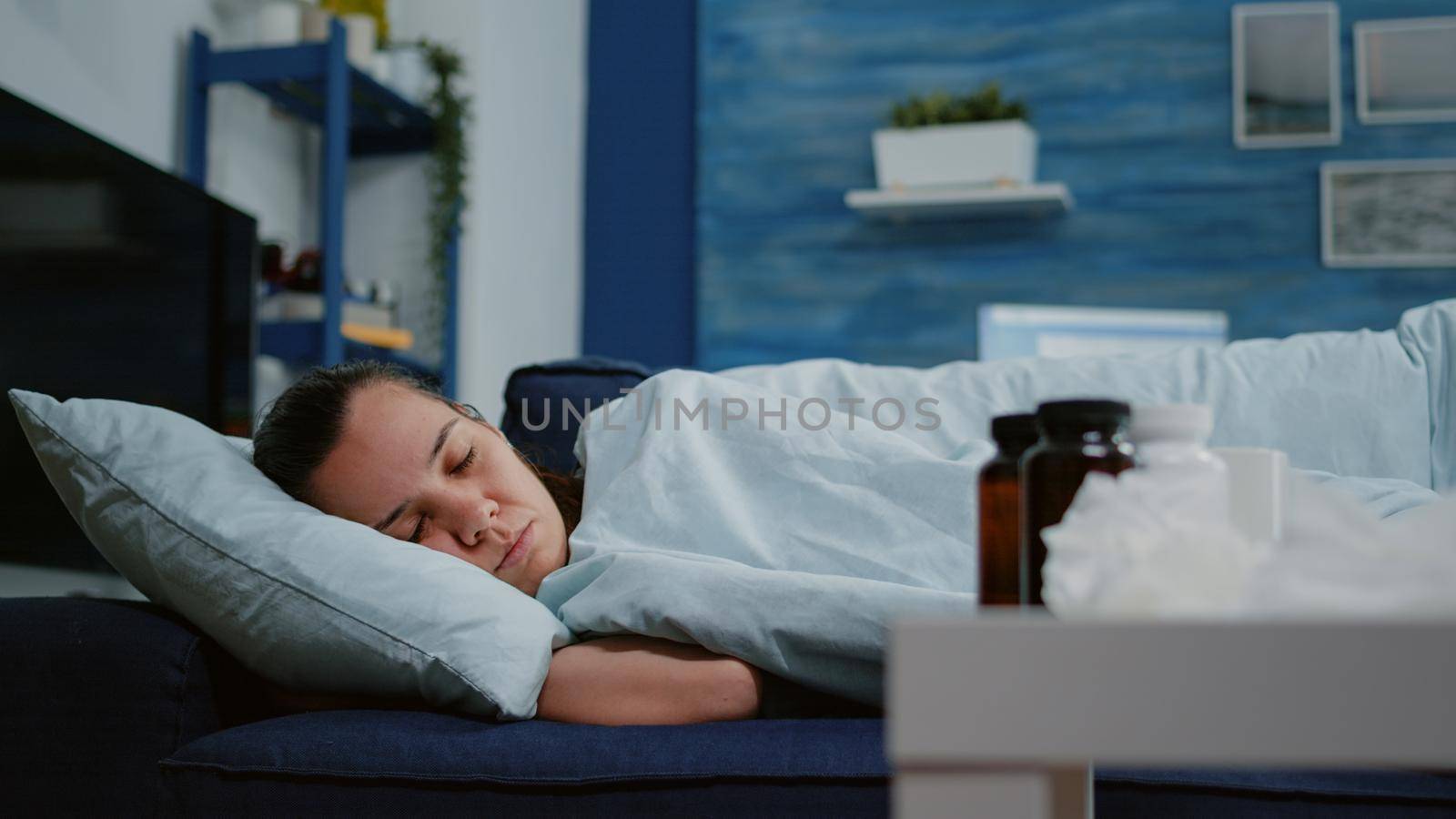 Close up of woman with disease sleeping in blanket on couch while having treatment against seasonal flu on coffee table. Sick adult resting with tissues, bottles of pills and medicaments.
