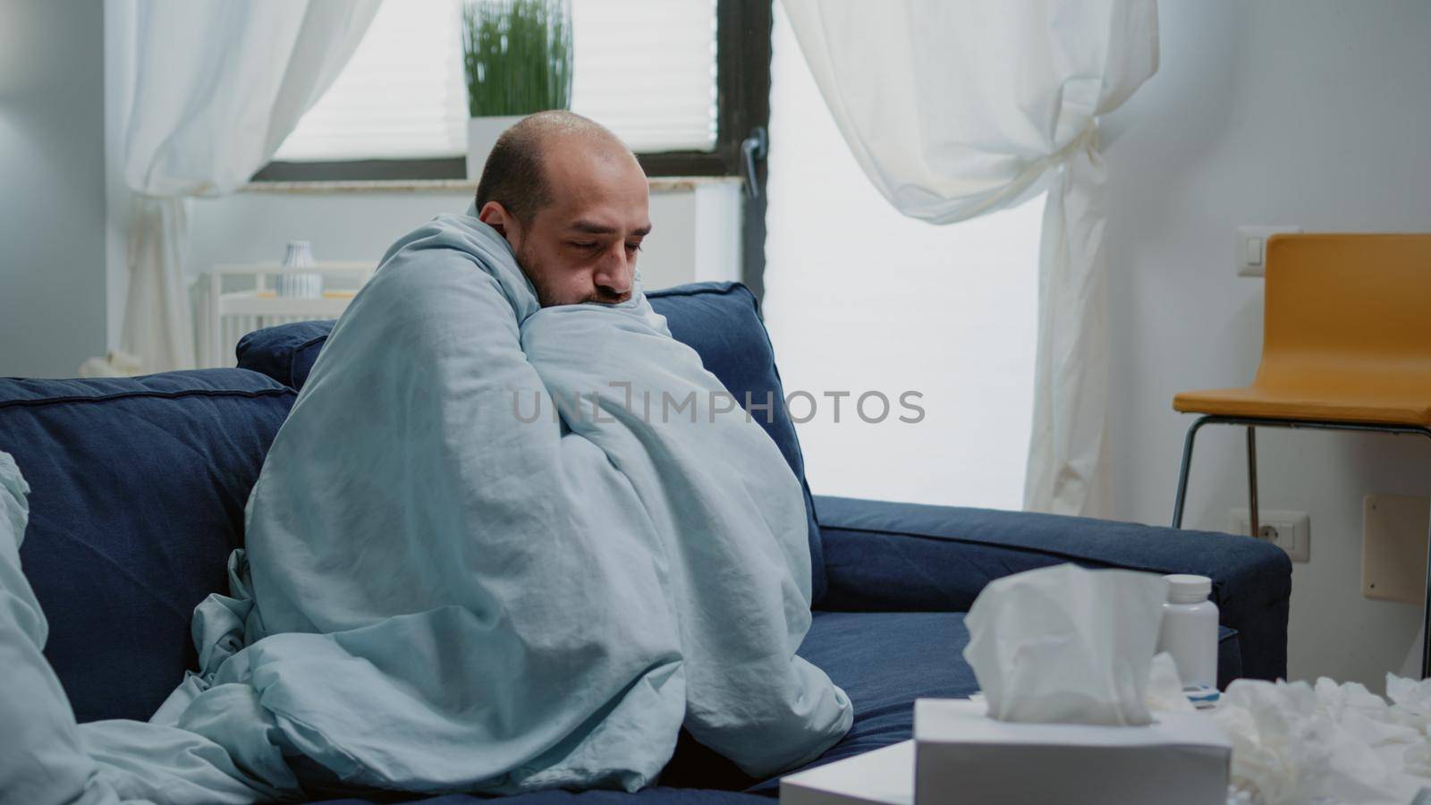 Person with flu feeling cold and shivering wrapped in blanket. Sick man having chills and fever symptoms while trying to get warm with cover, to cure illness with medicaments on table.