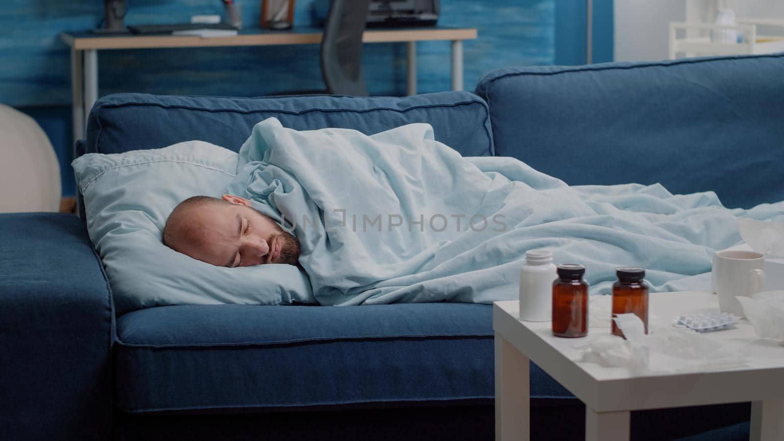 Unwell adult laying on sofa sleeping with medication on table. Sick man resting because of cold and flu, having bottles of pills, medicaments, tissues and cup of tea. Person with disease