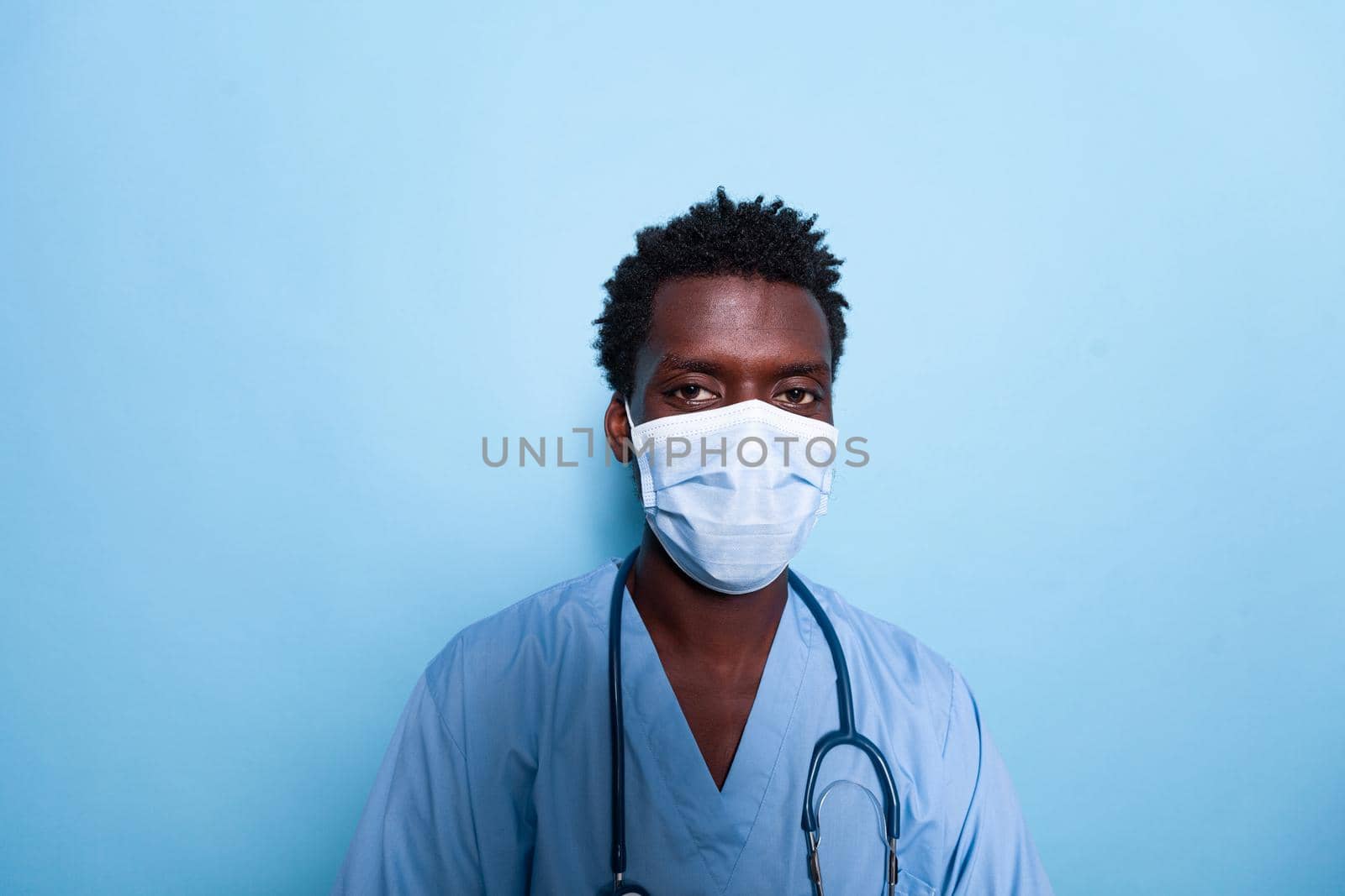 Portrait of medical assistant wearing face mask and uniform over blue background. African american healthcare nurse with stethoscope, having protection against covid 19 pandemic.