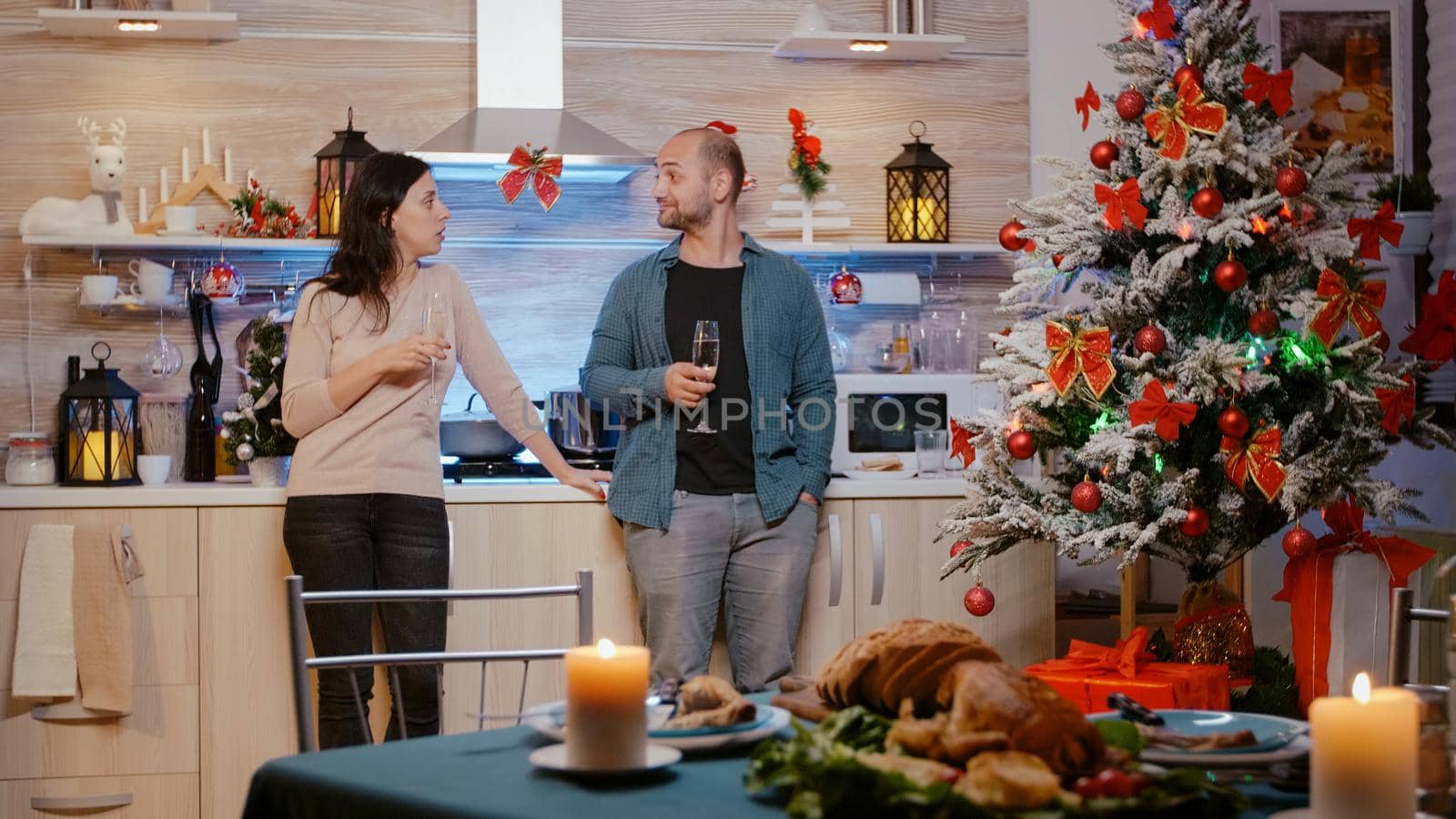 Festive couple standing and clinking glasses of champagne after enjoying christmas eve dinner. Man and woman chatting in decorated kitchen for seasonal holiday celebration. People celebrating