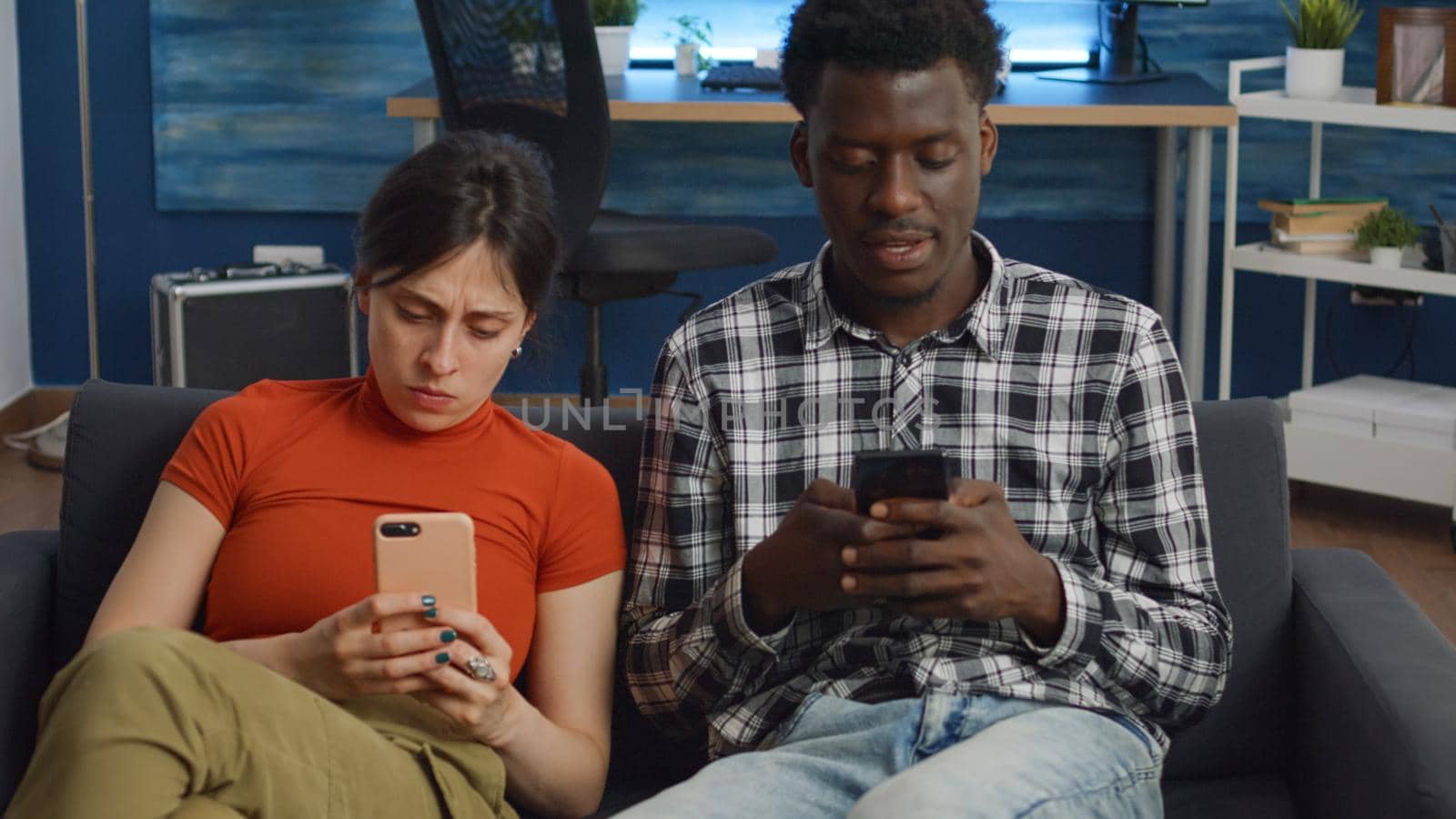 Interracial couple holding smartphones in living room on couch. Modern multi ethnic people with technology sitting together at home on sofa. Husband and wife using phone devices