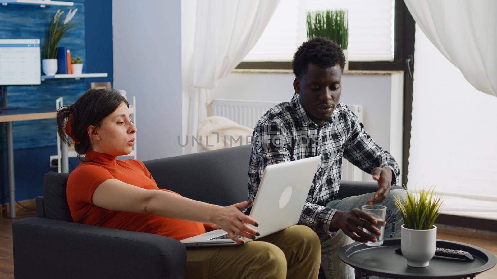 Interracial couple with pregnancy sitting together on couch while talking about child. Young pregnant woman using laptop and black man expecting baby and bonding in living room.