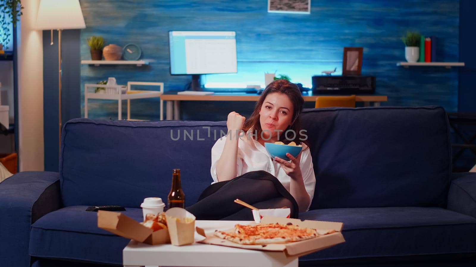POV of adult sitting on couch laughing while looking at TV and eating chips from bowl. Caucasian woman having fun watching television after work with takeaway food on table in living room
