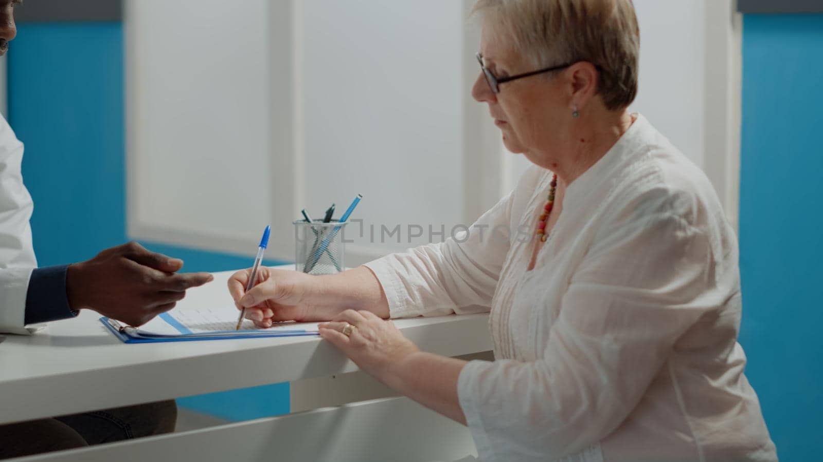 Close up of elder patient signing checkup documents with pen sitting at desk in medical cabinet. Young doctor needing signature on paper files from senior woman after healthcare appointment