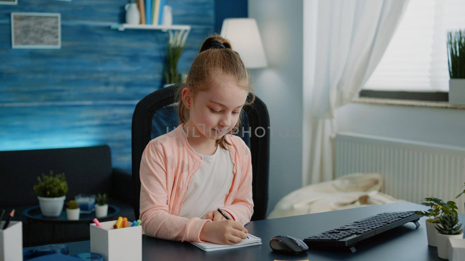 Schoolgirl writing on notebook with pen for homework. Little child concentrated on task for online remote lessons while sitting at desk with computer and textbook. Homeschooled pupil