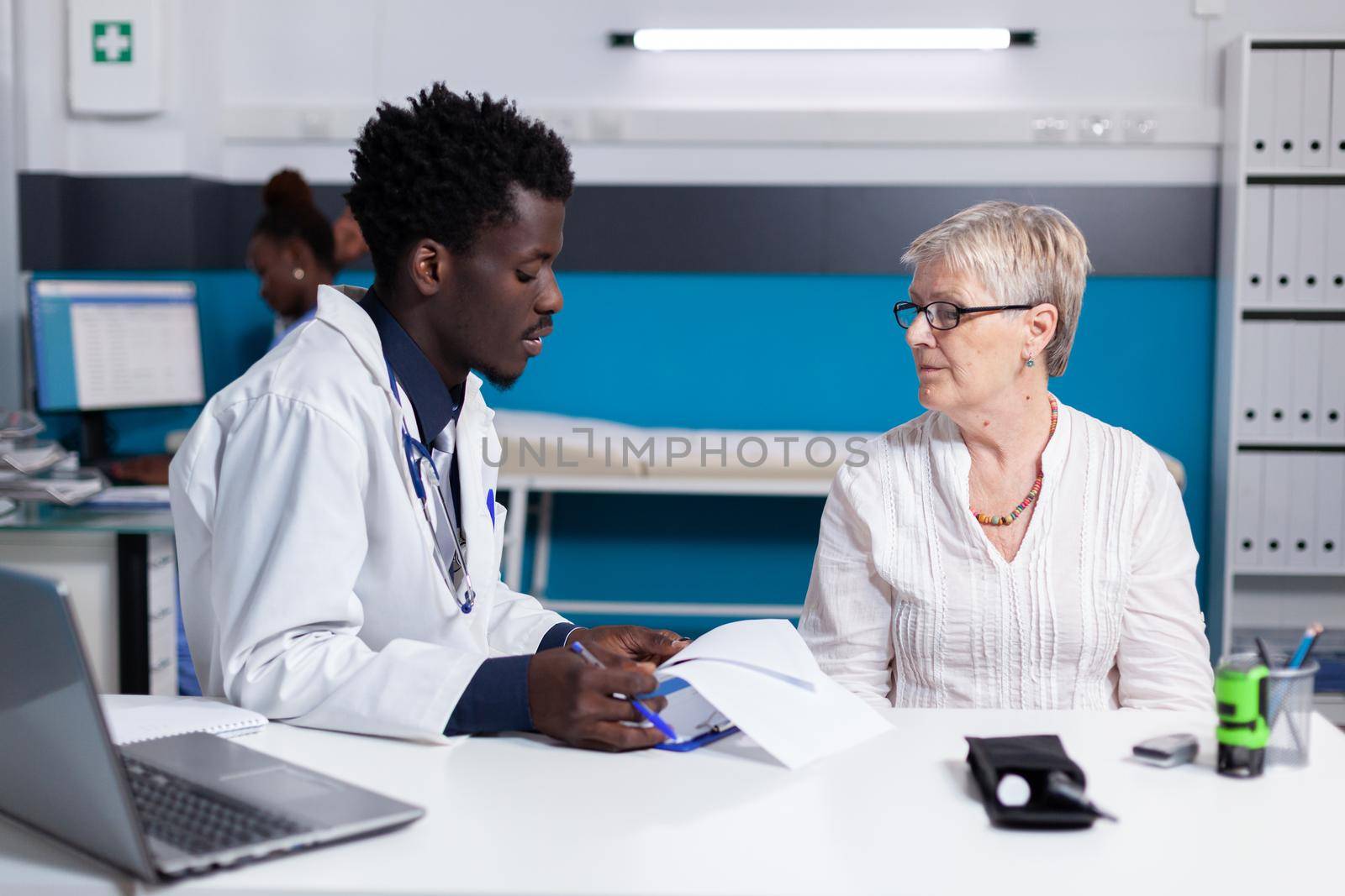 Black man with doctor profession talking to elder patient with disease in medical office. African american medic consulting senior woman about healthcare problems while sitting at desk