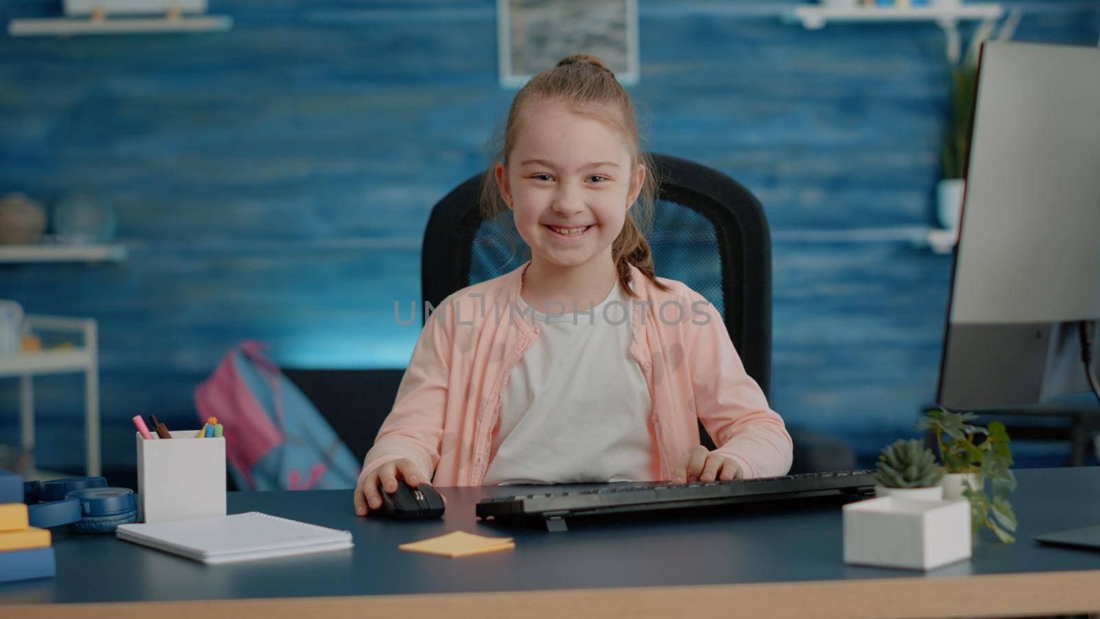 Portrait of schoolgirl at desk using computer and keyboard for online school courses and homework. Little child smiling and looking at camera while having technology for remote education