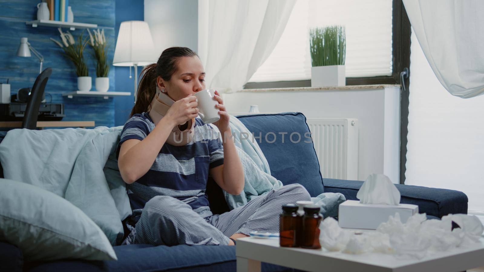 Adult with cold and flu wearing cervical neck collar while sitting on sofa. Ill woman with virus symptoms having vertebra injury and using neck foam for remedy, drinking cup of tea