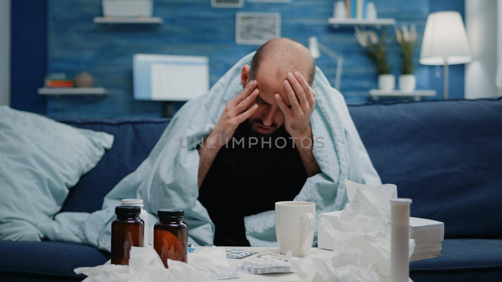 Close up of sick adult with headache rubbing temples in blanket. Man with migraine and flu symptoms trying to cure pain while having medicaments, pills, tea and thermometer on table