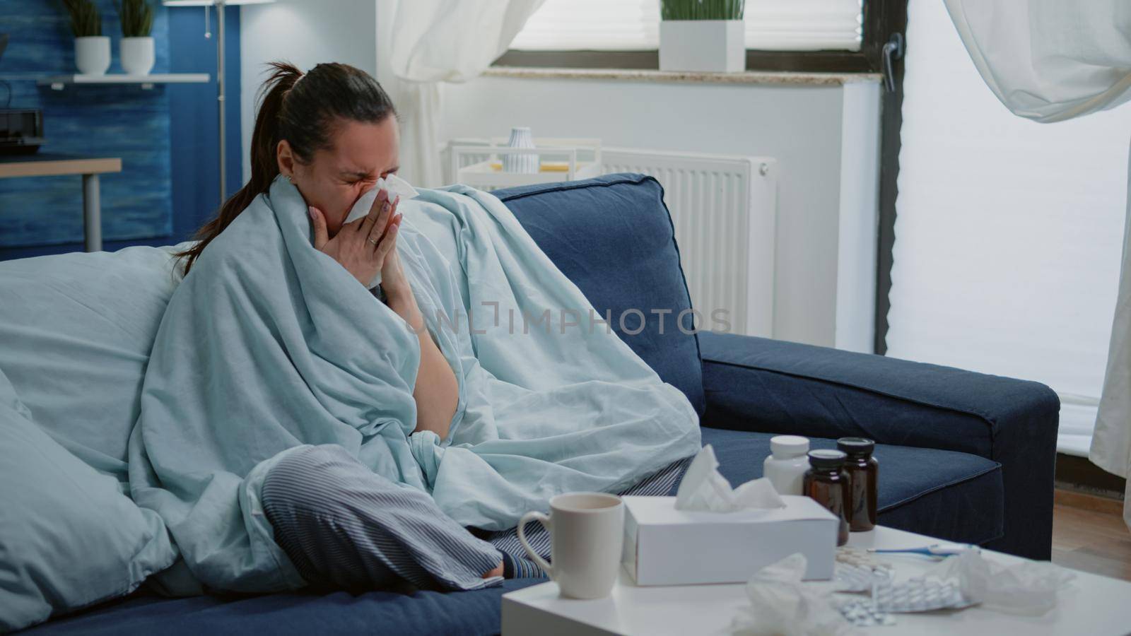 Person with cold blowing runny nose with tissue and sitting wrapped in blanket. Sick woman drinking cup of tea to cure sore throat, having bottles of pills for disease treatment.