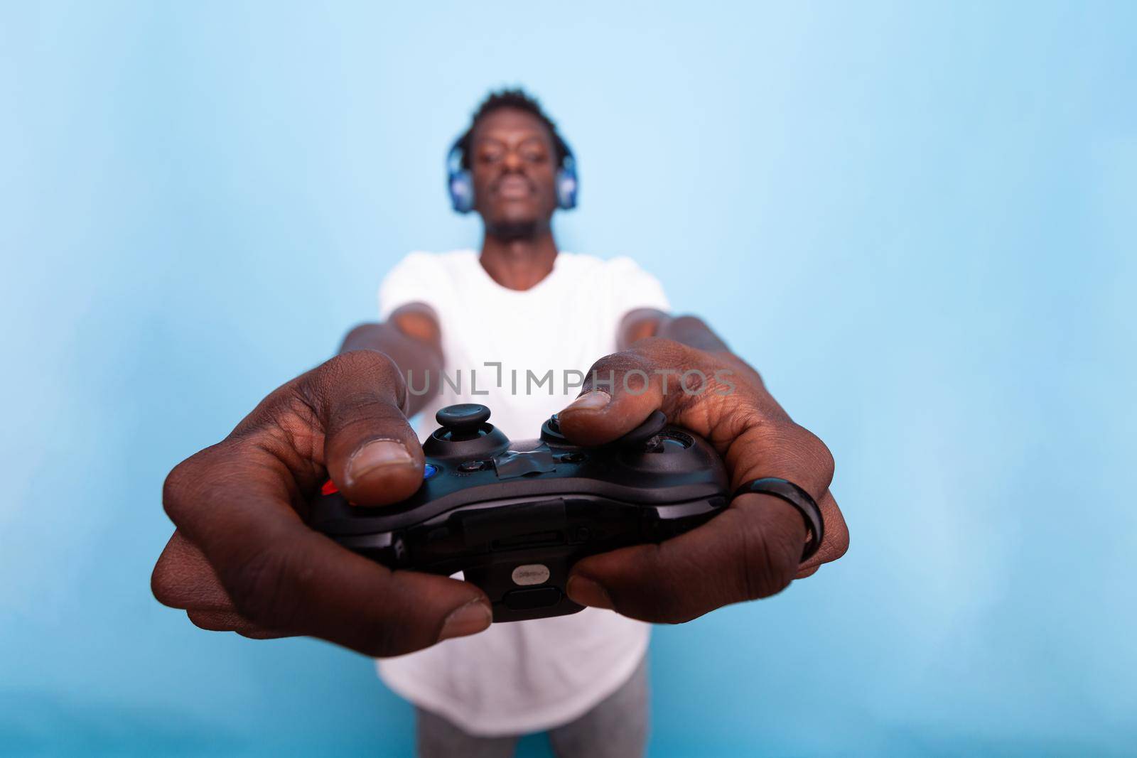 Close up of video games controller in hands of man. Person holding joystick at camera lens and wearing headphones. Gamer using gadget for online play, standing over blue background