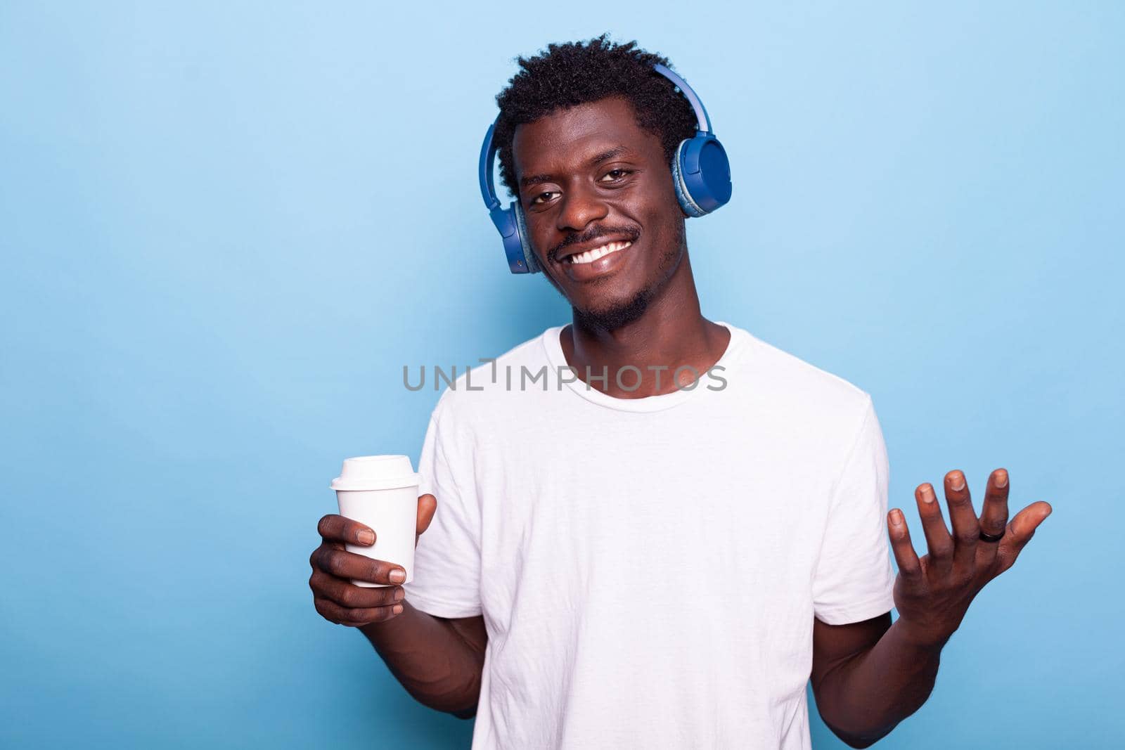 Man wearing headphones while holding cup of coffee and looking at camera. Young adult listening to music with takeaway drink in hand. Person feeling happy enjoying beverage and technology.