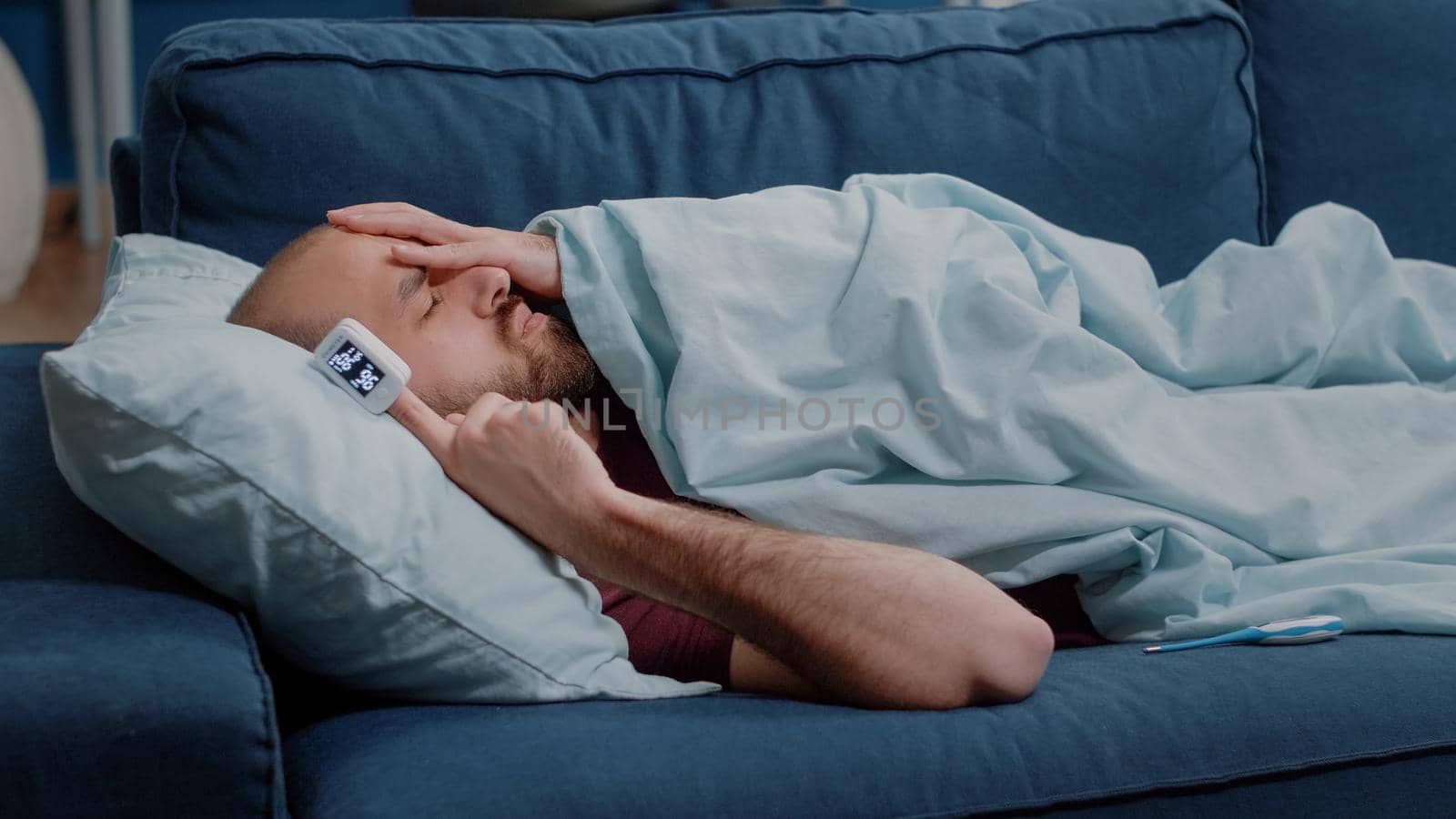 Sick adult using oximeter to measure raising oxygen saturation and pulse pressure. Ill person resting on sofa checking symptoms to treat sickness. Man with disease laying with blanket